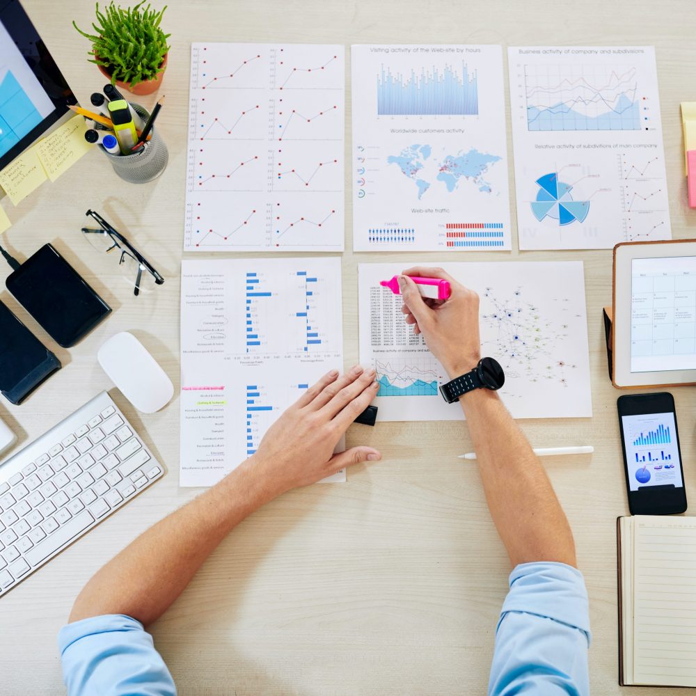 Hands of entrepreneur highlighting important figures in charts and financial reports, view from above